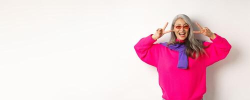 Fashion and beauty concept. Image of stylish asian senior woman in sunglasses smiling, showing peace signs and looking happy, standing over white background photo