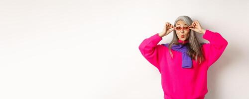 Trendy asian senior woman in sunglasses, pucker lips and looking surprised at camera, staring amazed at special offer, standing over white background photo
