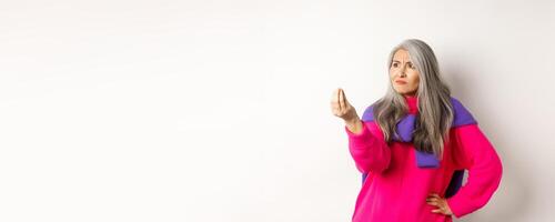 Grumpy asian grandmother complaining, showing italian what do you want gesture, shaking fingers and staring angry left at copy space, standing over white background photo