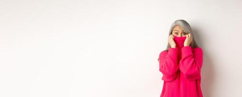 Portrait of funny asian grandmother hiding face in sweater collar, peeking at camera silly, standing over white background photo