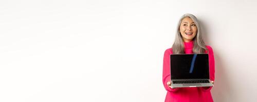 E-commerce concept. Happy beautiful asian senior woman with grey hair, looking aside and smiling, showing laptop black screen, standing over white background photo