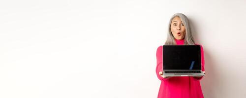 E-commerce concept. Impressed asian senior woman showing blank laptop screen and staring amazed at camera, standing over white background photo
