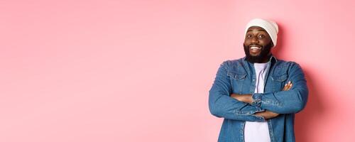 Happy Black man with beard, wearing beanie and denim shirt, looking intrigued and amused at camera, smiling with arms crossed on chest, pink background photo