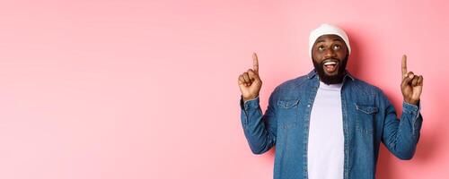 Happy african american man showing advertisement, smiling amazed and staring at camera, pointing fingers up, pink background photo