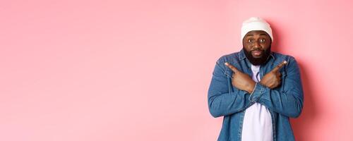 Confused Black man asking help with choice, pointing sideways and staring at camera, making decision, standing over pink background photo