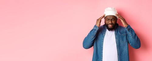 Distressed Black man having headache, touching head and grimacing with concerned face, standing over pink background photo