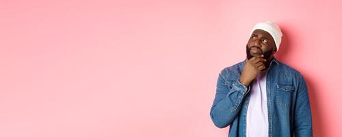 Image of puzzled and concerned black man, staring at upper left corner and thinking, making decision with doubts, standing over pink background photo