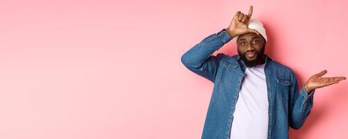 Sad african-american man showing loser sign on forehead and staring at camera, standing over pink background photo