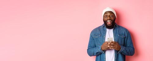 Technology and online shopping concept. Surprised young Black man using mobile phone, looking at camera amazed and happy after reading message, pink background photo