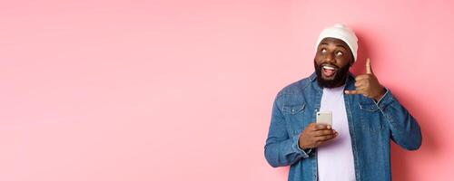 Happy Black man showing call me sign, making phone gesture and smiling, holding smartphone, standing in beanie and denim shirt over pink background photo