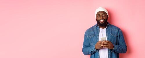 Handsome african-american guy using smartphone, smiling at camera, standing over pink background photo