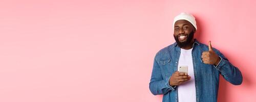 Technology and online shopping concept. Happy african-american guy showing thumb-up in approval while using mobile phone, standing satisfied over pink background photo