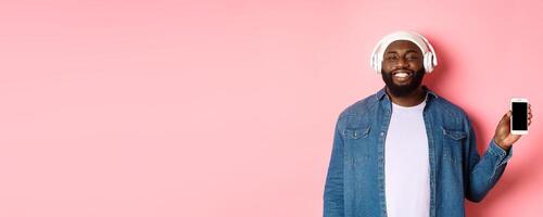 Happy hipster guy listening music on headphones and showing mobile screen, smiling satisfied, standing over pink background photo