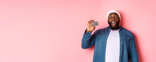Shopping concept. Amazed Black guy showing credit card, staring at camera impressed, recommending bank, standing over pink background photo