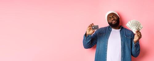 Shopping concept. Sad and whining Black guy showing credit card and dollars, grimacing reluctant, standing over pink background photo
