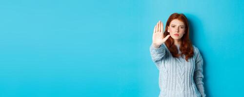 Serious and confident redhead girl telling to stop, saying no, showing extended palm to prohibit action, standing over blue background photo