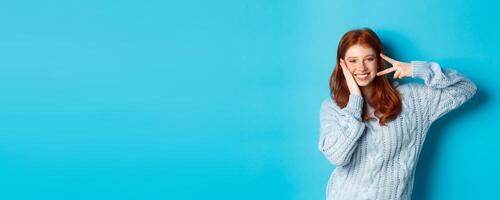 Cheerful redhead female model sending good vibes, smiling and showing peace sign, standing over blue background photo