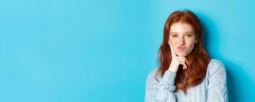 Close-up of beautiful redhead girl thinking, pucker lips and staring thoughtfula t camera, making choice, standing over blue background photo