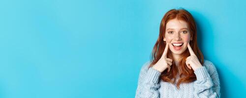 Close-up of cheerful teenage girl with red hair and freckles, poking cheeks, showing dimples and smiling with white teeth, standing over blue background photo