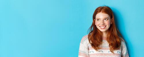 Close-up of beautiful redhead female model in winter sweater, smiling and looking left at copy space, standing over blue background photo