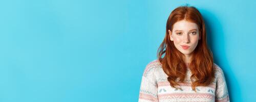 Close-up of redhead cunning girl having an idea, smirk and stare at camera satisfied with plan, standing over blue background photo