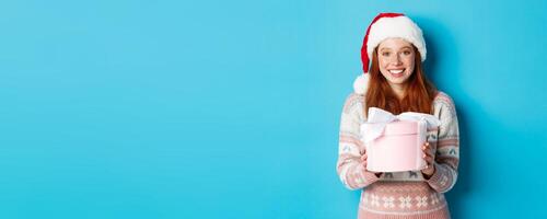 Winter and celebration concept. Adorable redhead girl giving you a present, wishing merry christmas, standing i santa hat against blue background photo