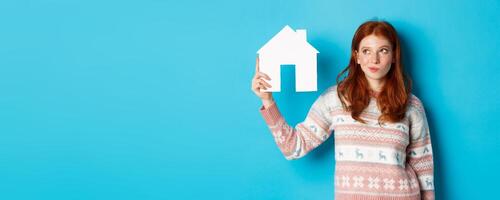 Real estate concept. Image of thoughtful redhead girl showing paper house model and thinking, searching for home or flat, standing against blue background photo