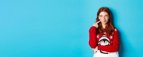 Winter holidays and Christmas Eve concept. Pretty redhead girl in xmas sweater, touching cheek thoughtful and smiling, making choice, looking at upper left corner and thinking, blue background photo