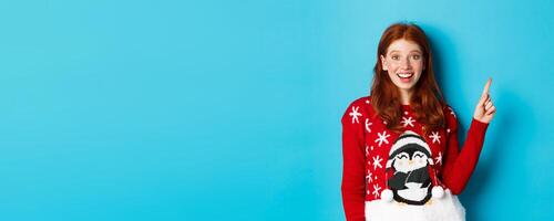 Merry Christmas. Cheerful redhead girl in xmas sweater, pointing finger at upper right corner and smiling excited, showing new year promo photo
