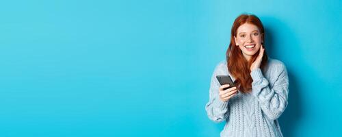 Beautiful redhead girl in sweater, smiling at camera, using mobile phone app, standing with smartphone against blue background photo