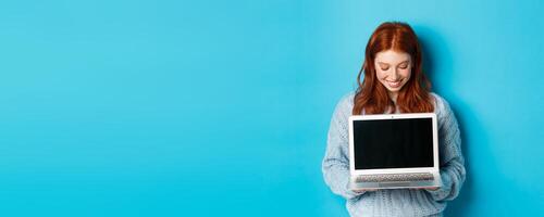 Cute redhead woman in sweater, showing and looking at laptop screen with pleased smile, demonstrating something online, standing over blue background photo