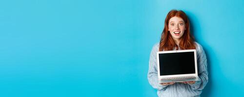 Excited redhead female freelancer showing laptop screen, staring at camera amazed, standing with computer against blue background photo