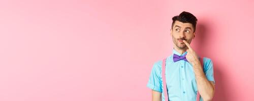 Image of handsome funny man in bow-tie looking thoughtful left and touching lip, thinking what to choose, making decision, standing over pink background photo