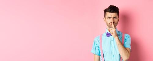 Angry and grumpy man in funny bow-tie shushing at camera, frowning and demand silence, tell to be quiet, scolding someone loud, standing on pink background photo