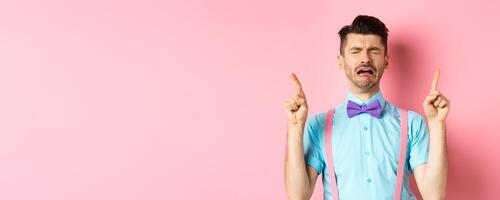 Sad and miserable guy sobbing and crying, pointing fingers up and something disappointed, standing upset on pink background photo