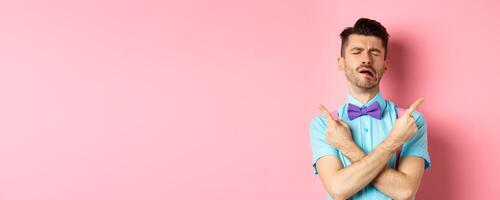 Sad and disappointed young man in bow-tie, crying and pointing fingers sideways, showing two things he wants, sobbing from unrequited love, standing on pink background photo