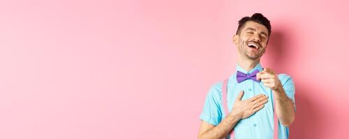 Handsome guy in bow-tie pointing finger at camera and laughing on something funny, checking out hilarious promo, standing on pink background photo