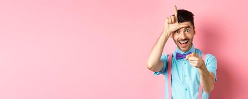 Image of funny guy pointing at you camera and showing loser sign, mocking person who lose, standing over pink background photo