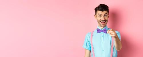 Cheerful smiling man in bow-tie pointing finger at camera, inviting or picking you, searching for candidate, standing over pink background photo