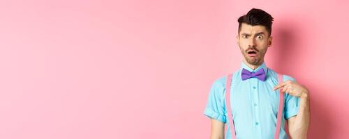 Image of confused funny guy in bow-tie and suspenders, pointing at himself as if being accused or chosen, raising eyebrow surprised, standing over pink background photo