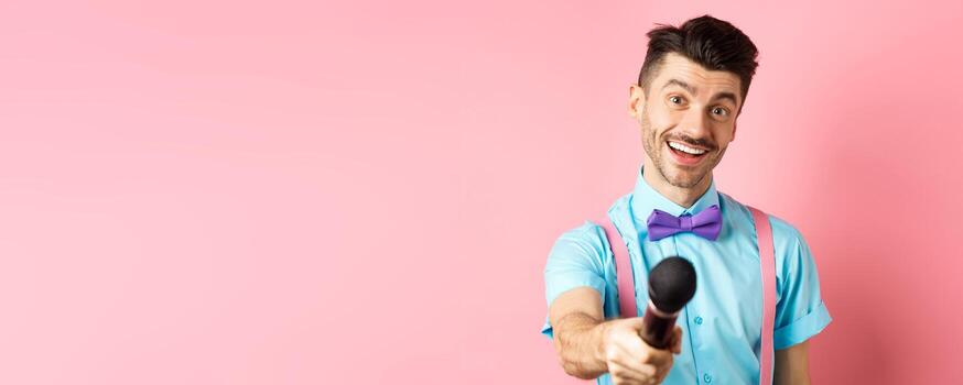 Smiling guy with moustache and bow-tie asking for comments or interview, stretch out hand with microphone, standing on pink background photo
