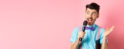 Image of male show host making speech, talking in microphone with clipboard under armpit, enteratin people on festive event, standing over pink background photo