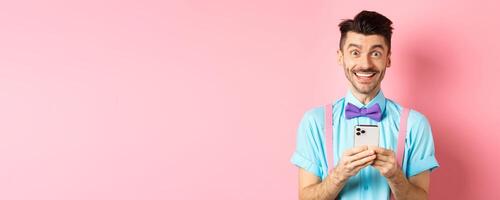 Online shopping. Happy guy in bow-tie, smiling at camera and using smartphone, standing on pink background photo