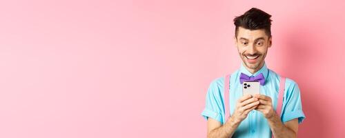 Online shopping. Happy guy looking at smartphone screen, reading message and smiling, standing on pink background photo