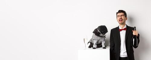 Sad man crying and drinking champagne from bottle while pug in cute party costume staring confused, standing over white background photo