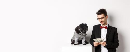 Handsome young man in suit standing near black pug in costume and counting money, working on parties, posing over white background photo