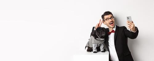 Cheerful dog owner in suit celebrating New Year with dog, taking selfie on smartphone near cute black pug in costume, white background photo