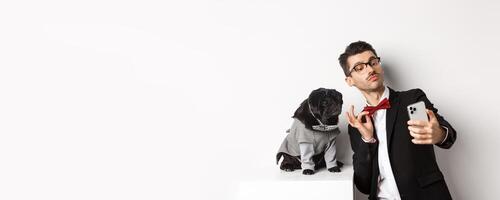 Cheerful dog owner in suit celebrating New Year with dog, taking selfie on smartphone near cute black pug in costume, white background photo