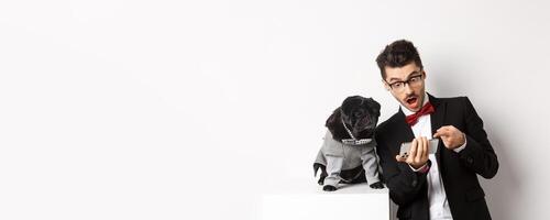 Amazed man showing to dog, pointing at mobile phone and look excited while pug sitting unbothered in costume, white background photo