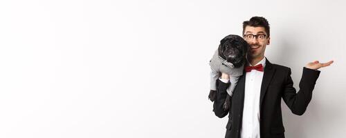 Handsome young man introducing pet product on hand, holding cute black dog on shoulder and smiling, showing something on white background photo